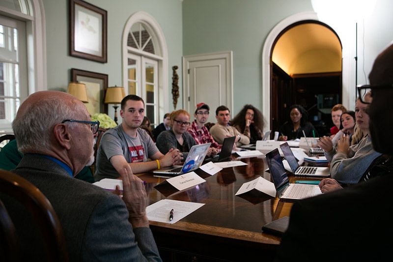 Group of students and 2 professors are seated around a dining room table. 