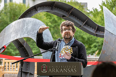 Professor holding paper models speaks in front of steel sculpture.