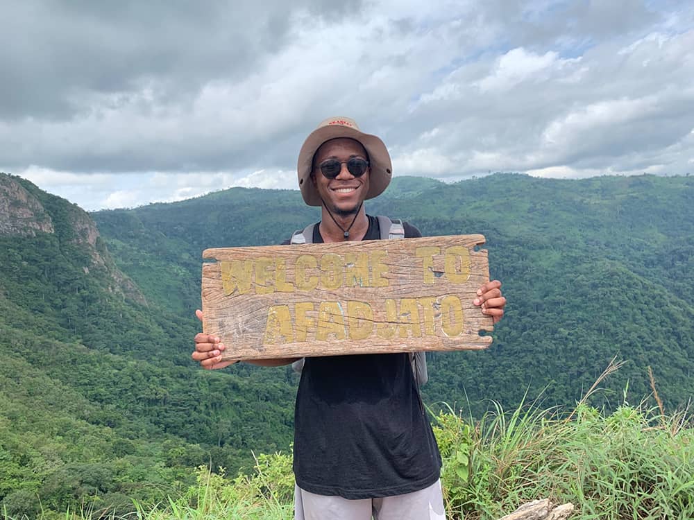 Young man holds sign at top of Mount Afadjato