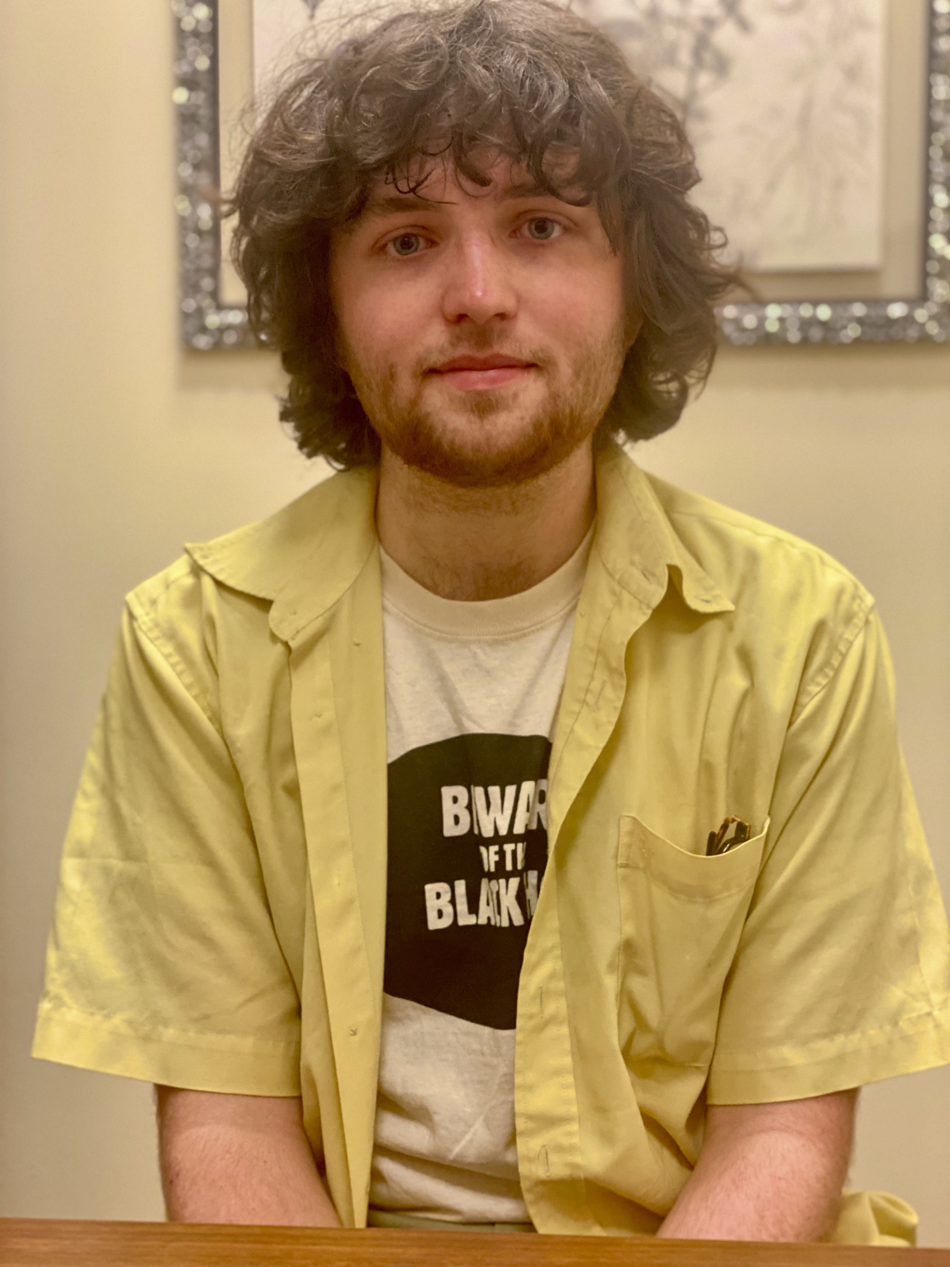 Ben sits and smiles into the camera. He's wearing a white t-shirt with black text and a yellow button-down shirt. His glasses are just visible in the front pocket of the button down.